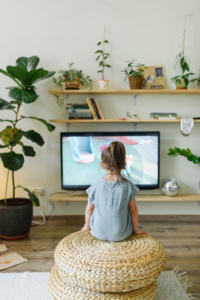 Back view of unrecognizable little kid watching television in living room with potted plants in house