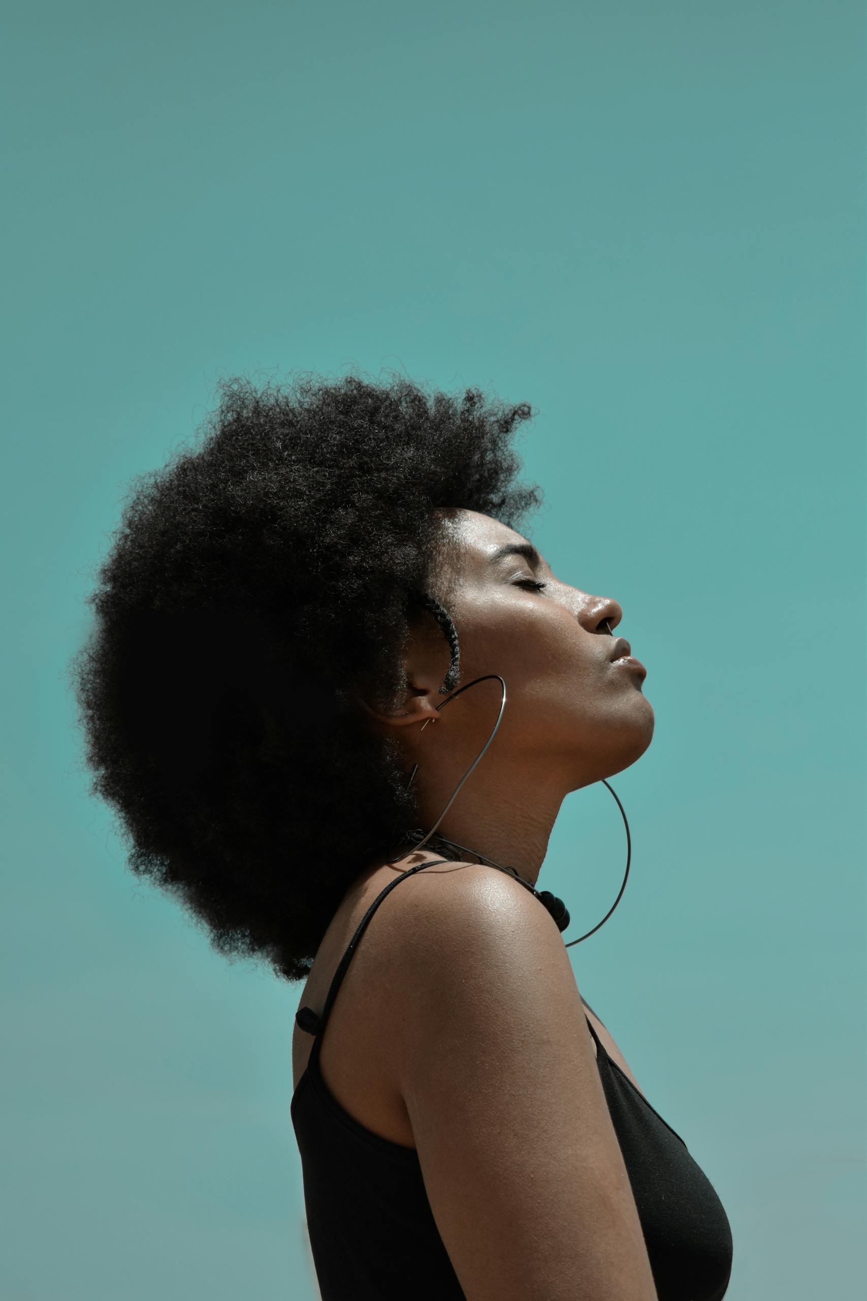 From below side view of young reflective ethnic female model with big earrings and closed eyes under blue sky