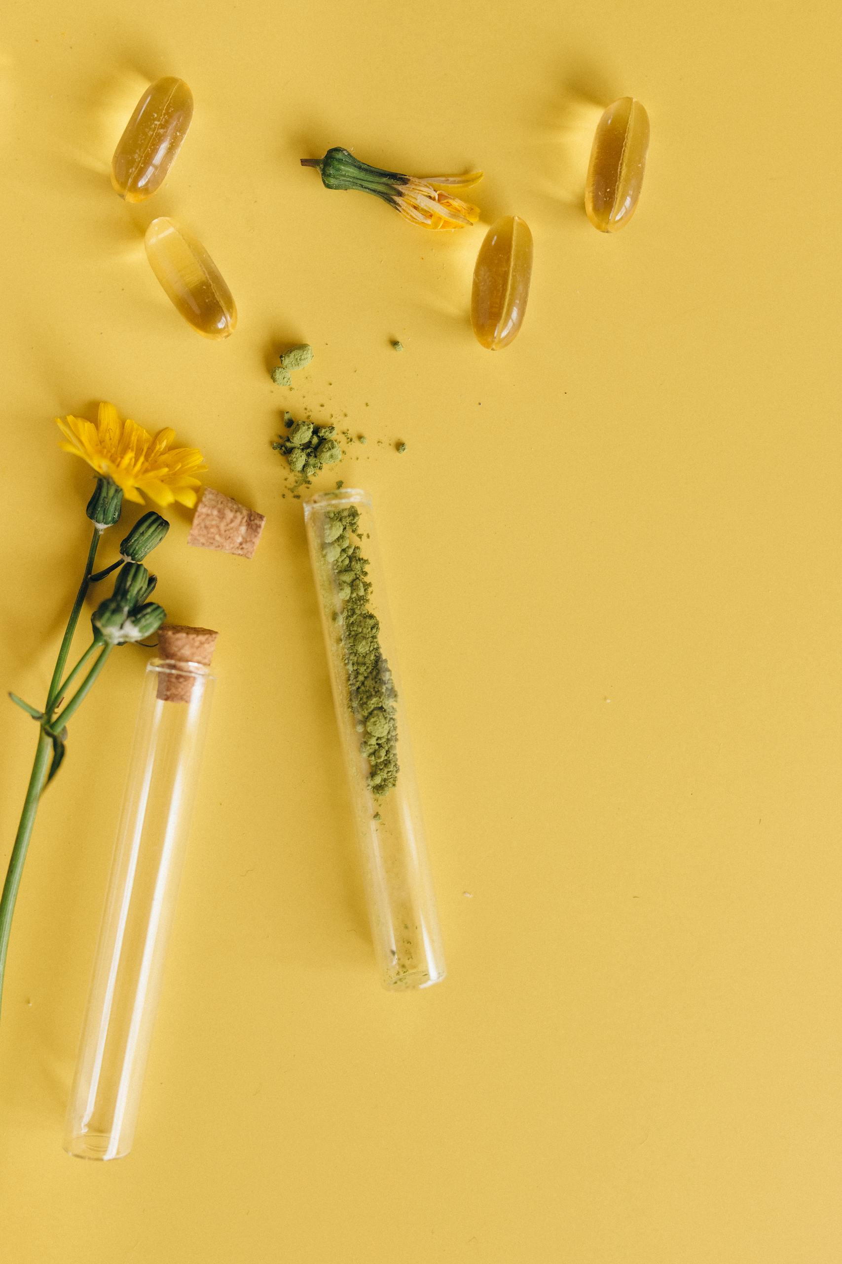 Top view of herbal capsules and flowers with test tubes on a vibrant yellow surface.