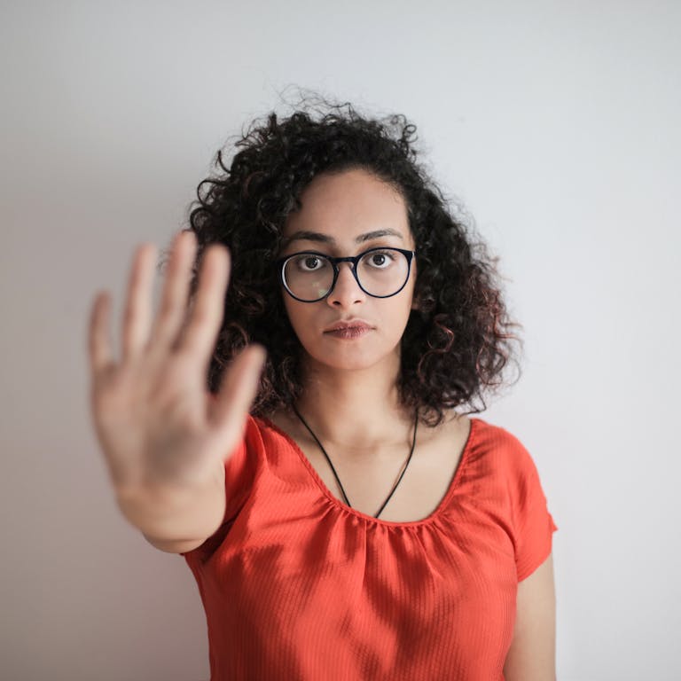 A serious woman with curly hair and glasses making a stop gesture against a white background.