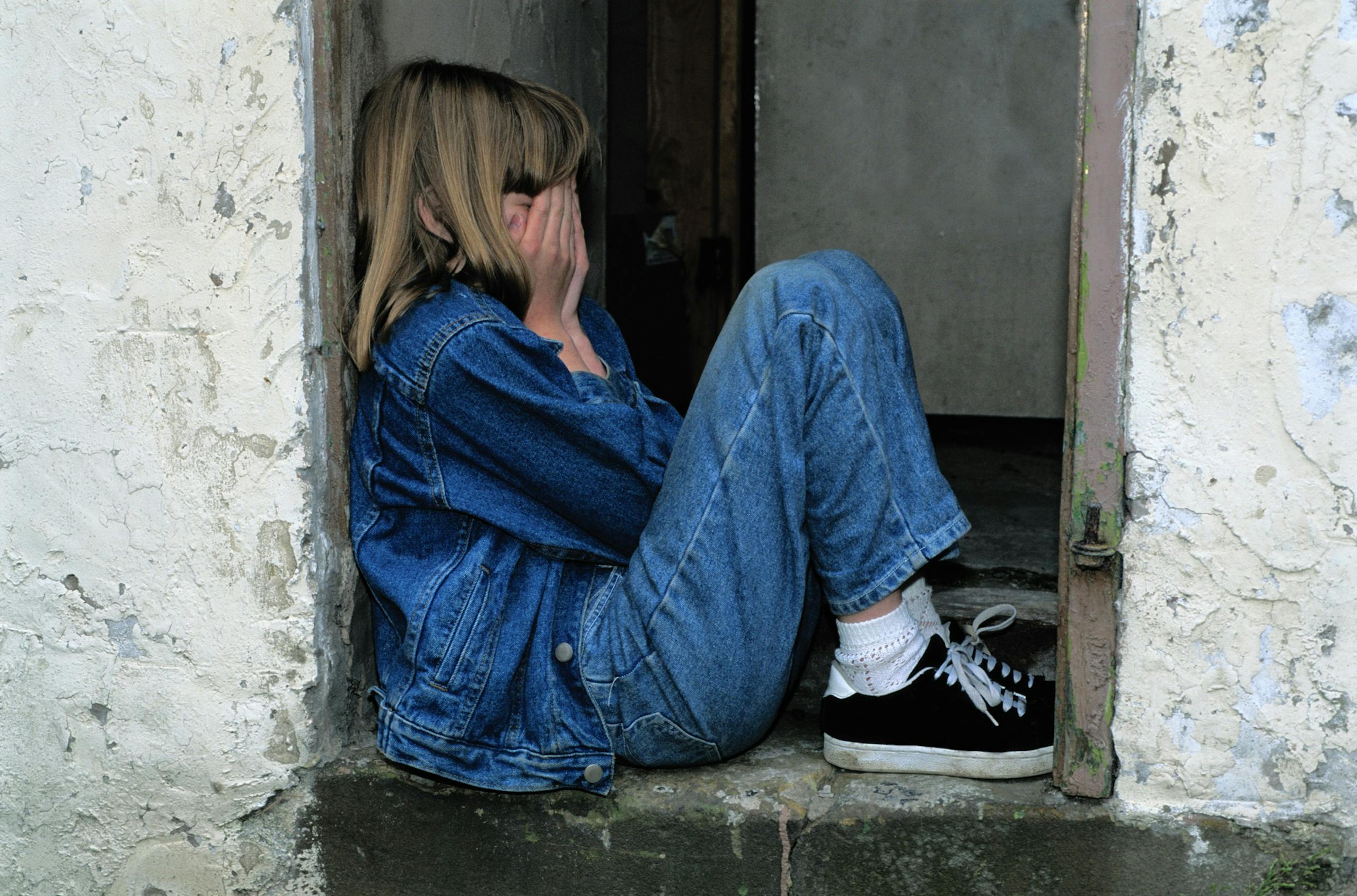 A young girl in denim sits alone in a doorway looking distressed, conveying loneliness.