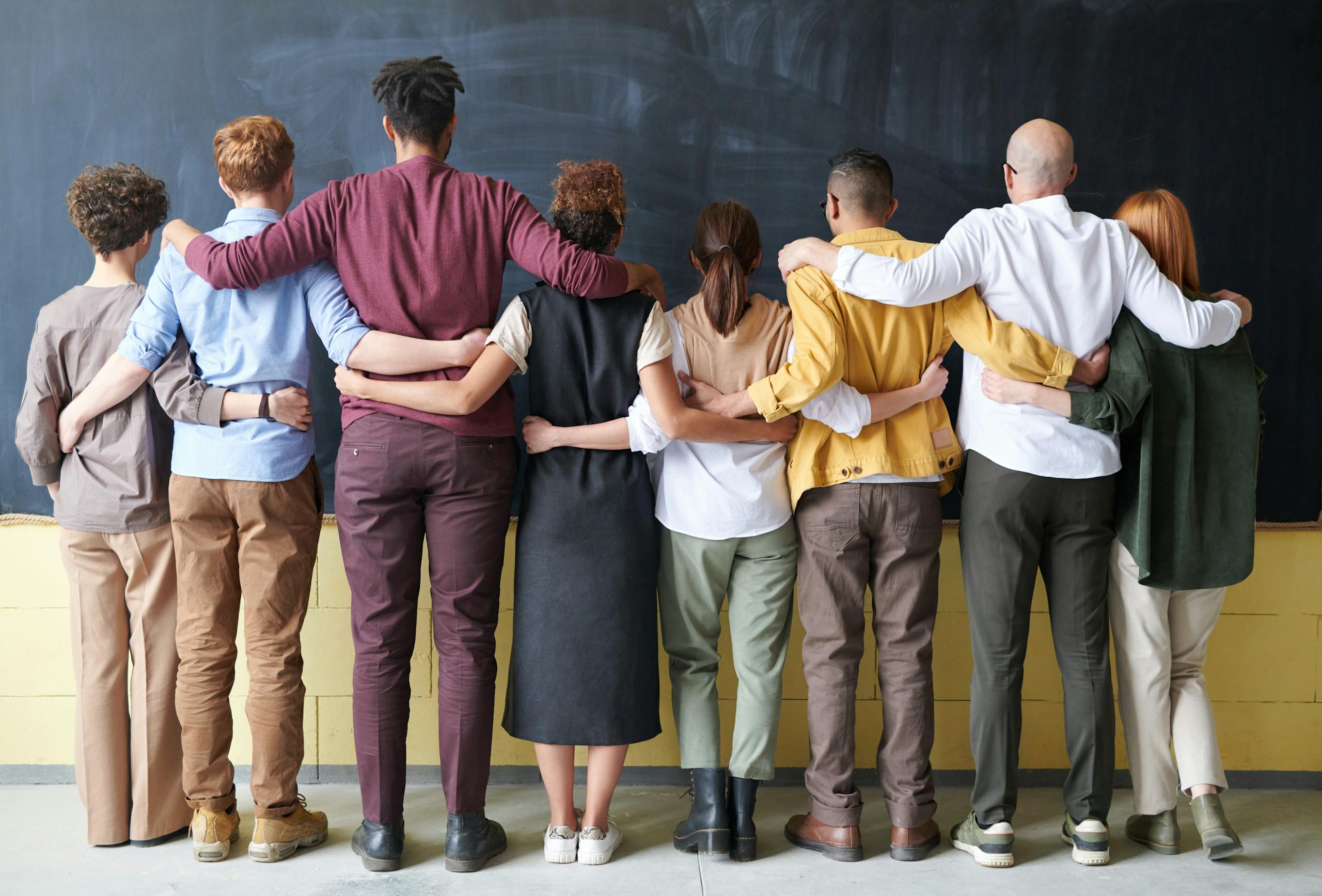 A diverse group of adults in casual outfits hugging in front of a chalkboard, symbolizing teamwork.