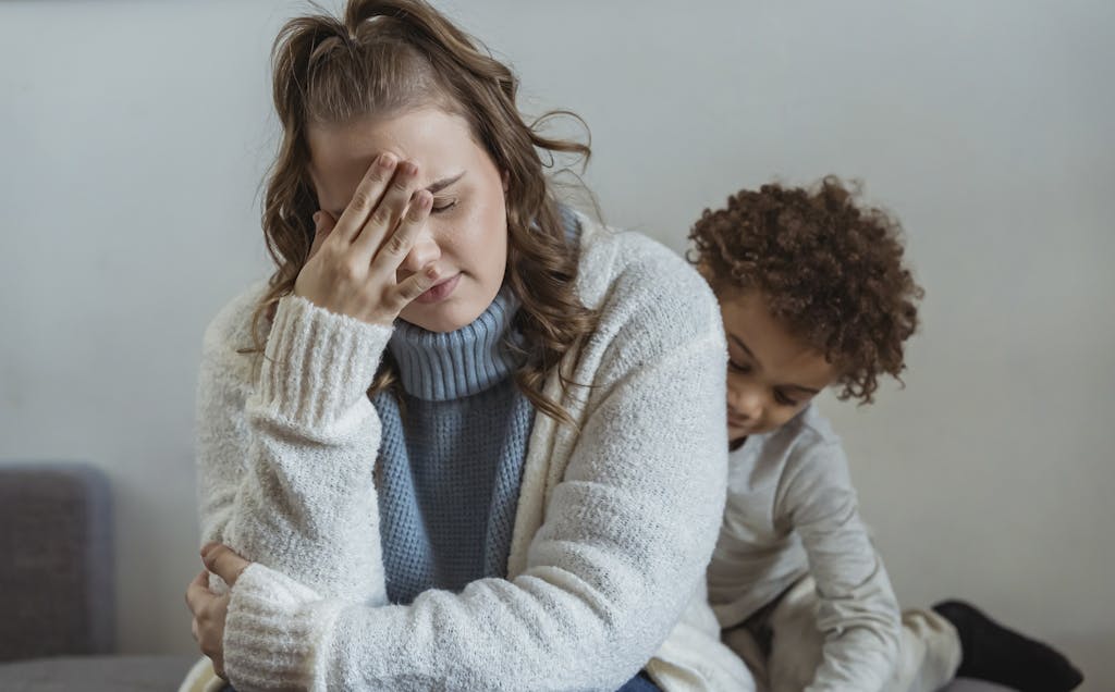 Frustrated mother with hand on forehead and closed eyes sitting near African American son near wall in room at home