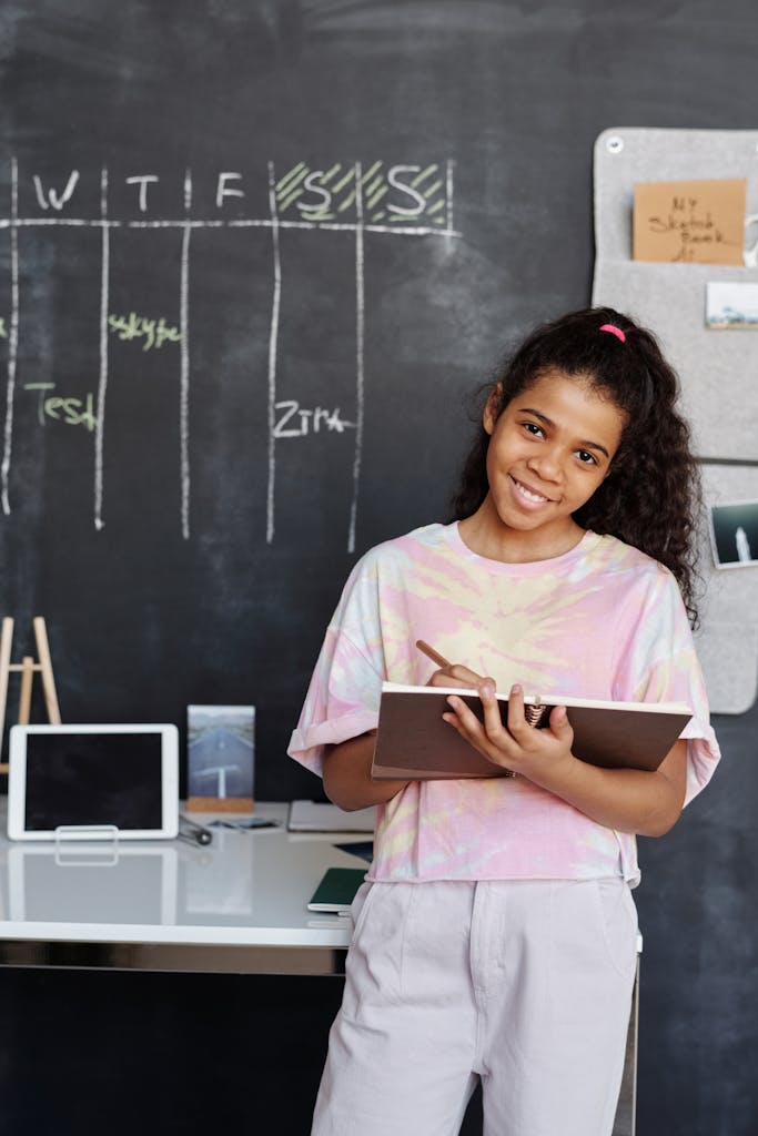 Teen girl smiles while studying indoors, holding a notebook in front of a blackboard.