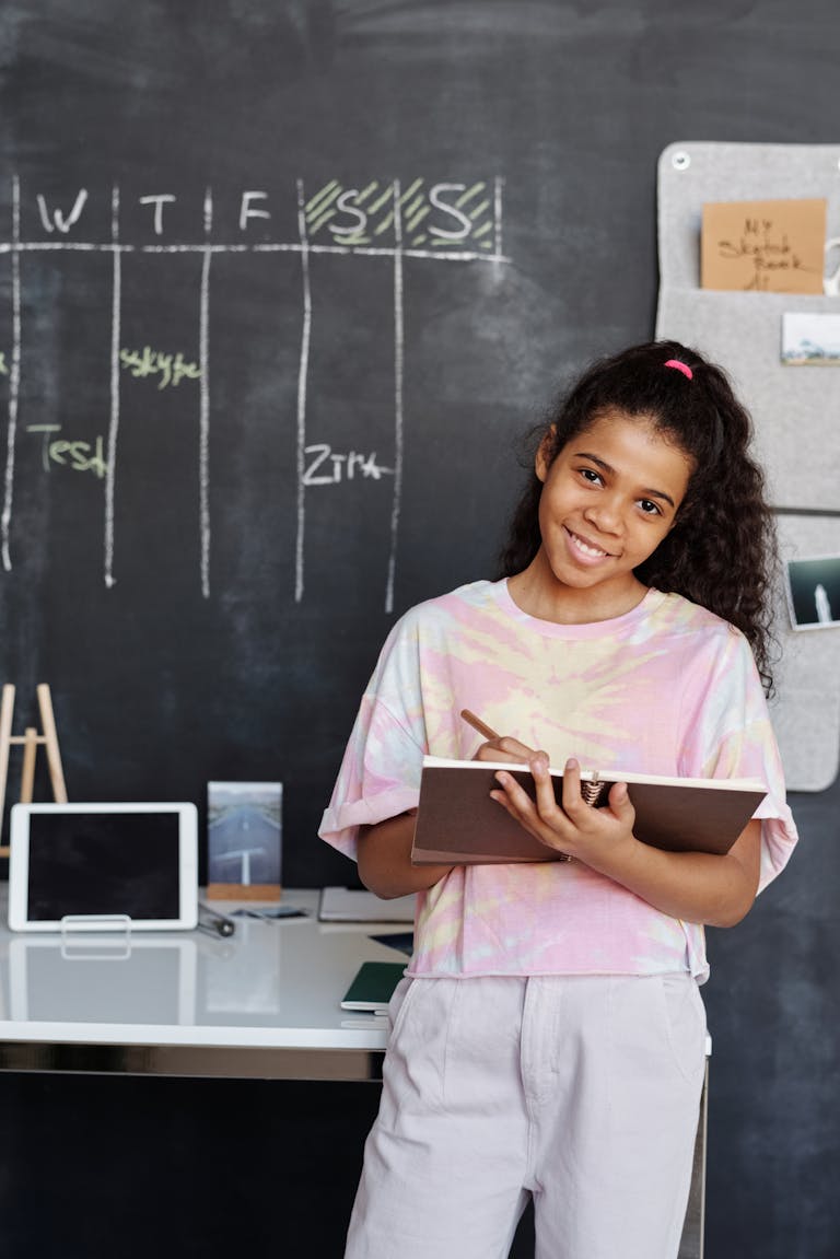 Teen girl smiles while studying indoors, holding a notebook in front of a blackboard.