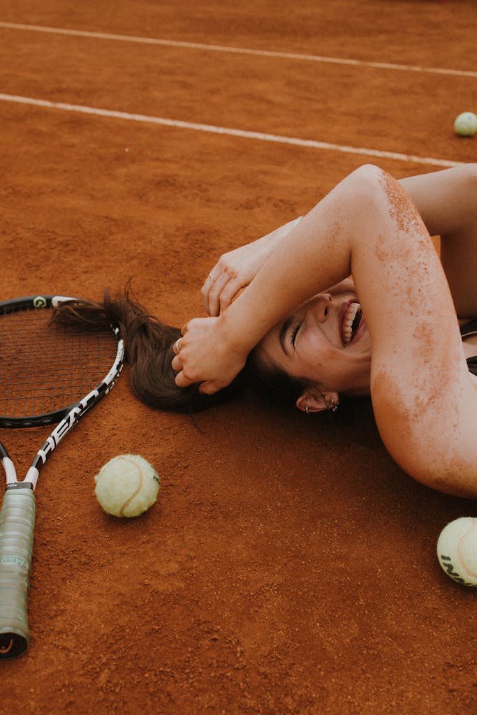 A young woman laughing and lying on a clay tennis court surrounded by tennis balls and racket.