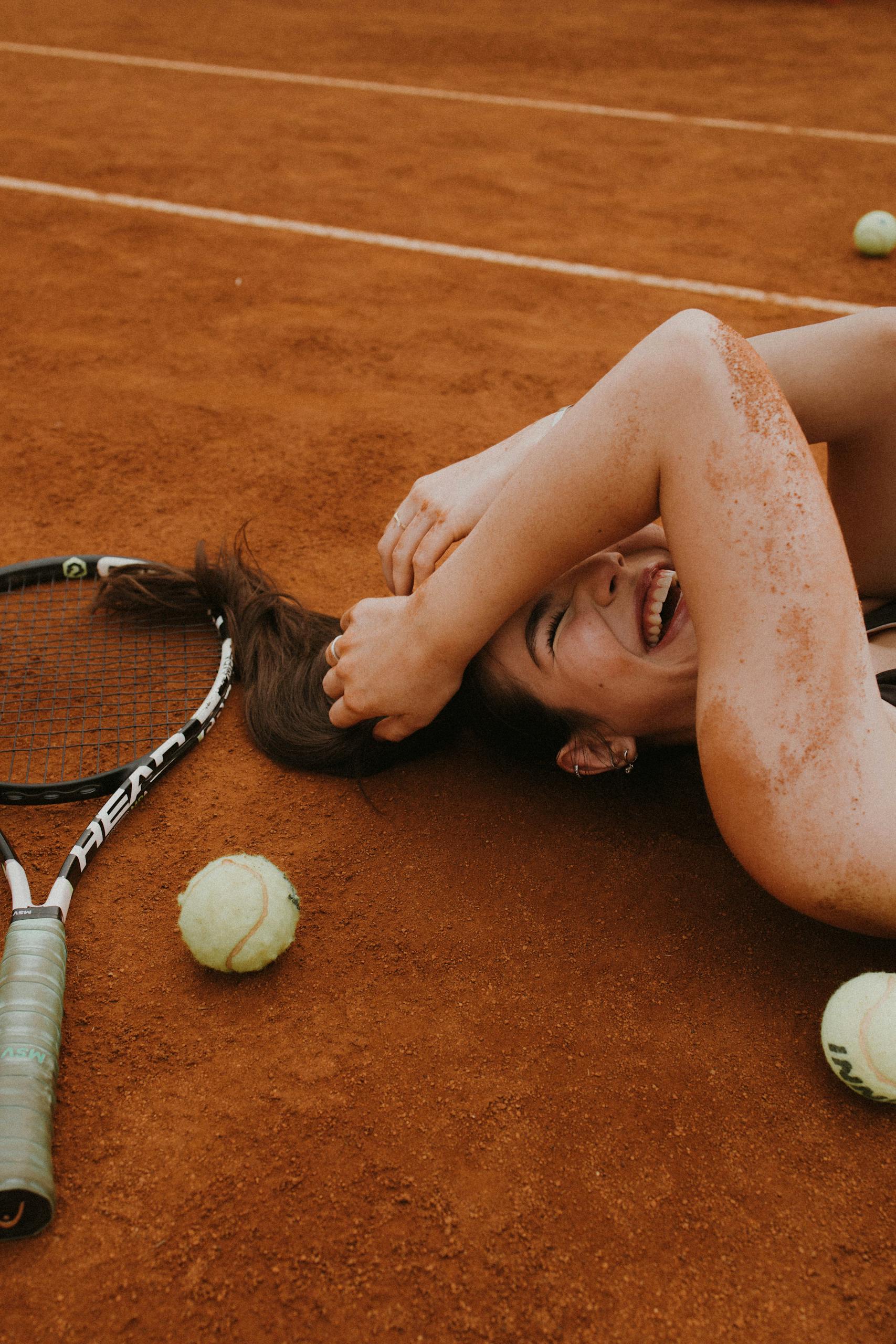 A young woman laughing and lying on a clay tennis court surrounded by tennis balls and racket.